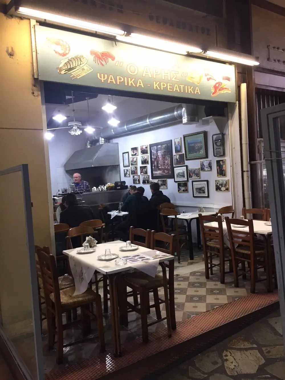A cozy interior of a taverna with wooden tables and chairs, covered with white tablecloths. The walls are adorned with framed pictures, and a sign above the entrance displays text in Greek along with images of seafood. People are sitting inside, and a staff member stands behind a counter.